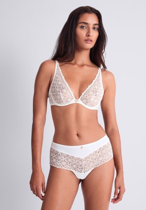 Femme aux longs cheveux foncés portant un ensemble de lingerie blanche en dentelle avec des motifs floraux, debout devant un fond gris clair uni.