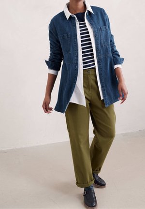 Persoon draagt een gelaagde outfit met een blauwe spijkerjack, wit overhemd, marineblauw gestreept topje, olijfgroene broek en zwarte schoenen, staand binnen.