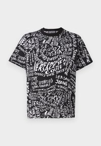 Juodas medvilnės marškinėliai su chaotišku dizainu, kuriame balta tekstas, įskaitant "Versace" ir "Jeans Couture", skirtingais dydžiais ir orientacijomis.