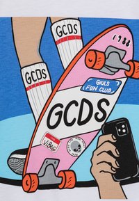 Rosa skateboard med orange hjul, "GCDS"-logotyp och klistermärken; blå bakgrund; vita strumpor med röda ränder; hand som håller en smartphone.