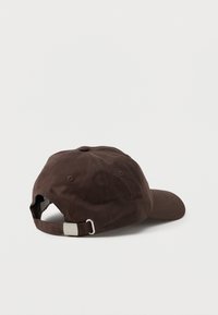 Casquette de baseball ajustable marron avec boucle en métal à l'arrière, posée sur une surface blanche unie, la visière tournée vers la droite.