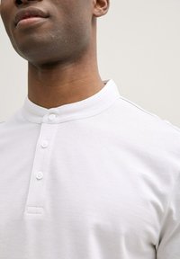 Witte henley-shirt van gestructureerde stof, met een staande kraag en een knoopsluiting met drie knopen, getoond op de bovenste torso van een persoon.