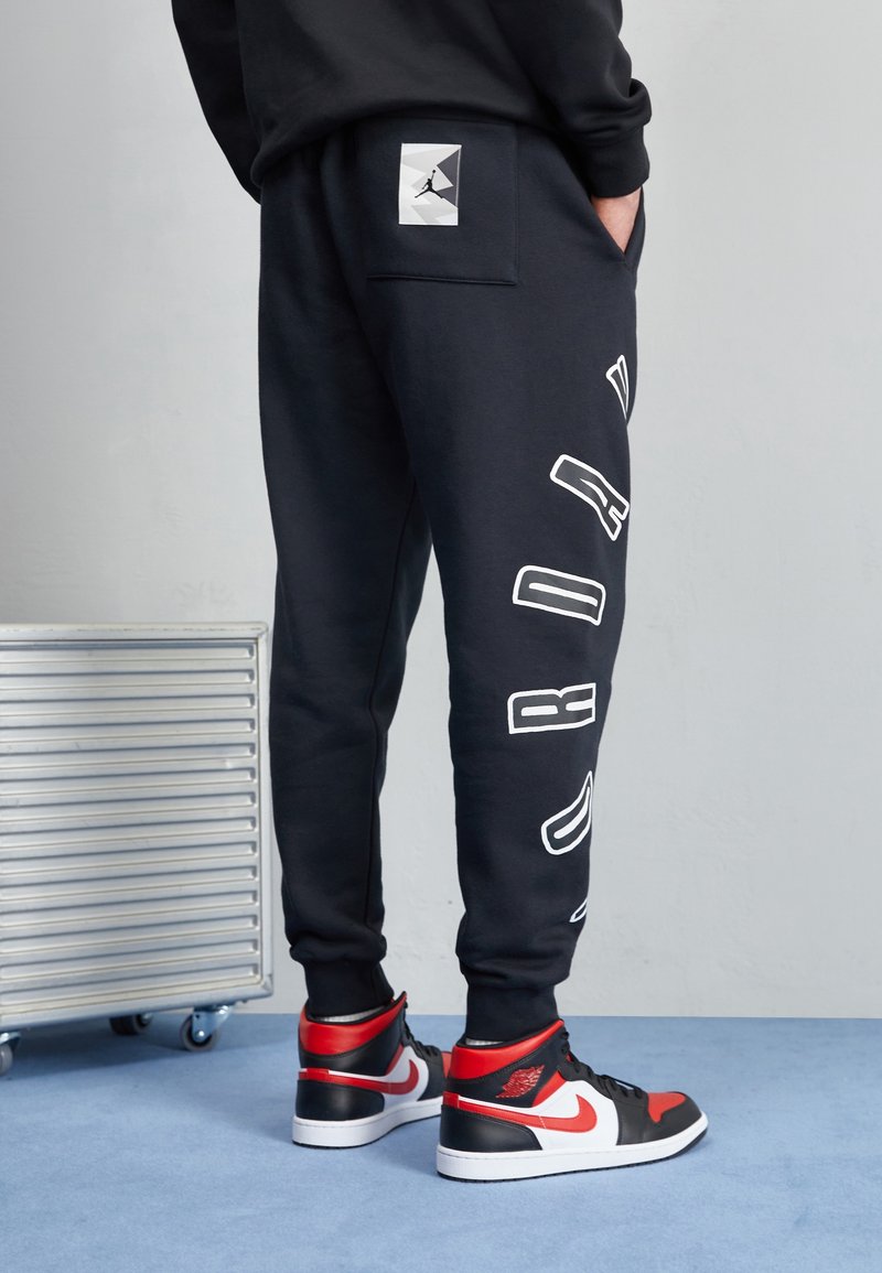 Svarta sweatpants i mjukt tyg med en stor vit logga på sidan och en fyrkantig loggficka. Kombinerade med svart-röda högskor.