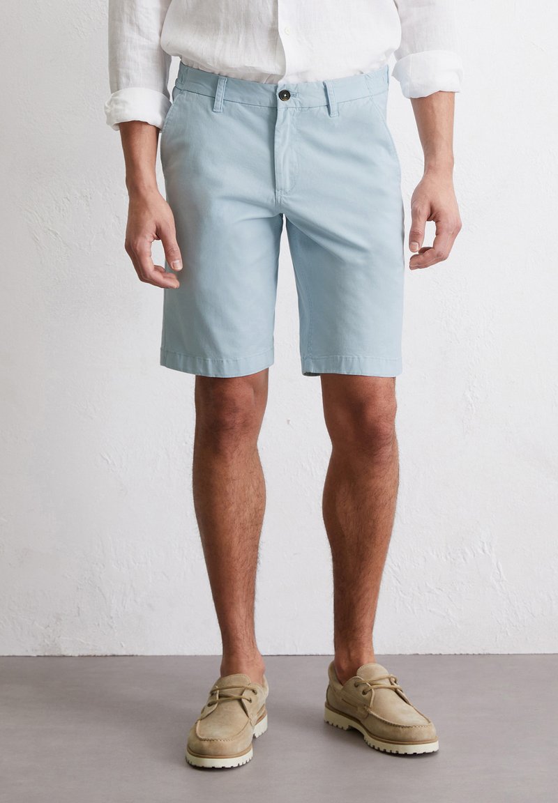 Shorts en coton bleu clair avec une taille plate, passant de ceinture, poches latérales et longueur au-dessus du genou, associés à des chaussures en daim beige.