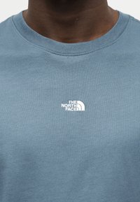 Κοντινό πλάνο ατόμου που φορά μπλε μπλούζα λαιμόκοψης με λευκό λογότυπο "The North Face" στο στήθος.