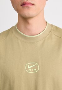Olivově zelené tričko Nike s kulatým výstřihem, na kterém je světle zelené logo s nápisem "AIR". Hladký materiál a krátké rukávy.