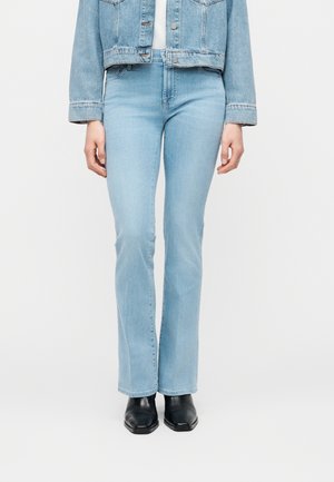 Személy világoskék bővülő fazonú farmert, fekete, négyzet alakú orrú csizmát és rövid, világoskék farmerdzsekit visel, fehér háttér előtt áll.