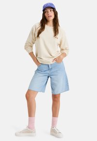 Beige sweatshirt met ronde hals, lichtblauwe denim shorts, roze ribbel sokken en witte sneakers. Blauwe pet achterstevoren gedragen.