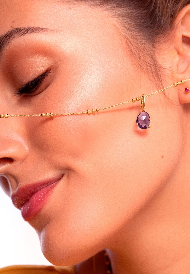 Une chaîne en or présente un pendentif en pierre précieuse violette, avec de petites perles métalliques le long de la longueur. Une peau lisse et une couleur de lèvres subtile sont visibles.