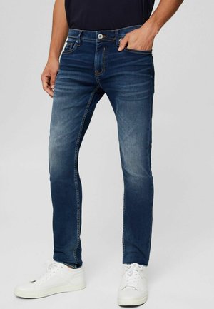 Mann trägt dunkelblaue Slim-Fit-Jeans und weiße Turnschuhe, steht mit einer Hand in der Tasche vor einem schlichten Hintergrund.
