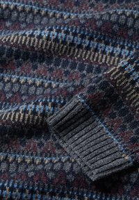 Tuvojoties vilnas džemperim, redzams piedurkne ar daudzkrāsainu Fair Isle rakstu, kurā ir tumši zila, pelēka, zila, bēša un bordo nokrāsas.