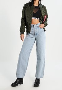 Zielona satynowa kurtka bomberka z zamkiem, czarny crop top z siateczki, jasnoniebieskie dżinsy o szerokich nogawkach oraz czarne botki na platformie. Czerwona metka na kurtce.