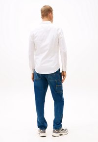 Camicia bianca a maniche lunghe con polsini bottonati, abbinata a pantaloni in denim blu con tasche e dettagli cuciti, indossati con sneakers grigie.