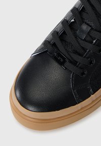 Svart syntetisk sneakersko med en slät yta, rund tå, svarta snören, gummisulor och diskreta varumärkesdetaljer.