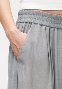 Χέρι που ακουμπάει μέσα στην τσέπη της ελαστικής μέσης ενός φαρδιού, ανοιχτόγκρι υφασμάτινου παντελονιού με λευκό τοπ ορατό από πάνω.