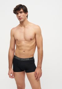 Млад мъж с къса тъмна коса, облечен с черни боксерки Tommy Hilfiger, стои на неутрален светъл фон.