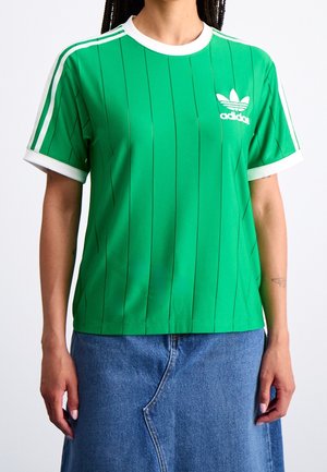 Personne portant un t-shirt vert Adidas avec des bandes blanches et un logo, associé à une jupe en jean bleue sur un fond clair uni.