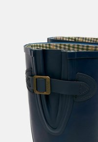 Sötétkék gumicsizma texturált felülettel, arany csattal díszítve, zöld és fehér kockás béleléssel.