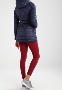 Chaqueta acolchada azul marino con capucha, mallas rojas y zapatillas deportivas grises. Presenta un diseño acolchado y un corte de longitud media.