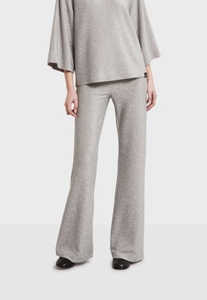 Donna che indossa pantaloni argentati larghi e abbinati con top ampio e scarpe nere, in piedi su sfondo uniforme.