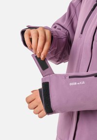Lilla vandtæt jakke med sort inderside og Velcro-justerbare manchetter. Har en lynlås foran og trykt branding.