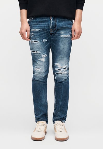 Μπλε τζιν τζιν με φθαρμένα σκισίματα και ξεθωριασμένες κηλίδες, στενή εφαρμογή, με κλασικό σχεδιασμό πέντε τσεπών. Συνδυασμένο με ανοιχτόχρωμα αθλητικά παπούτσια.