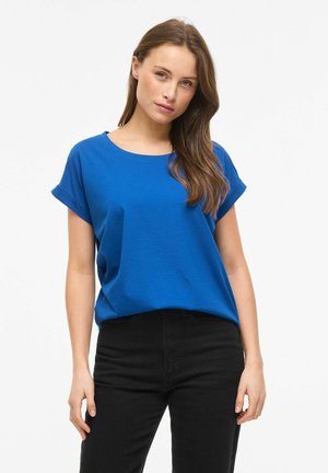 Femme aux longs cheveux bruns portant un t-shirt bleu à manches courtes et un pantalon noir, debout devant un fond blanc uni.