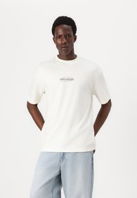 Camiseta de algodón blanca con mangas cortas y un escote redondo, que presenta un pequeño logo gris "HOLLISTER" centrado en el pecho.