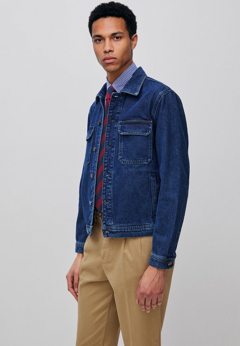 Jonge man die een blauw spijkerjack draagt over een geruit overhemd met een rood-blauw gestreepte stropdas en beige broek, staand tegen een effen achtergrond.