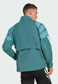 Teal sportska jakna s visokim ovratnikom, s mrežastim panelima, bijelim detaljima i slobodnim krojem straga za ventilaciju.