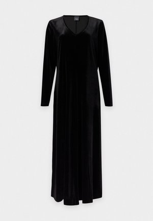 Femme aux cheveux clairs portant une longue robe en velours noire et des talons noirs, debout avec une main sur la hanche sur un fond blanc uni.