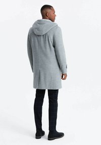 Abrigo gris con capucha de tejido texturizado, de corte largo, con costuras laterales y un ajuste relajado, combinado con pantalones de cuadros oscuros y botas negras hasta el tobillo.