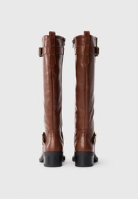 Une paire de bottes en cuir marron hautes avec des talons noirs, des boucles près du haut et de la cheville, vues de l'arrière sur un fond uni.
