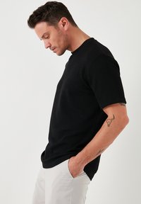 Schwarzes, geripptes Kurzarm-T-Shirt mit rundem Halsausschnitt, getragen mit Händen in den Taschen, kombiniert mit hellen Hosen.