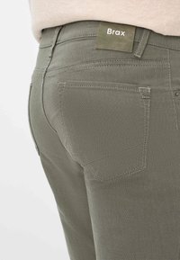 Jean en denim Brax vert olive avec une surface texturée, une poche arrière avec un logo discret et des détails de couture renforcée.