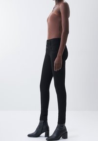 Jeans ajustados negros con una cintura alta y cierre de dos botones, combinados con una camiseta sin mangas de canalé marrón y botas negras hasta el tobillo con tacón apilado.
