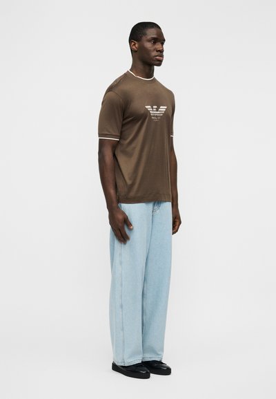 Καφέ βαμβακερό t-shirt με λευκή επένδυση και λογότυπο στο στήθος, σε συνδυασμό με ανοιχτό μπλε τζιν παντελόνι φαρδιάς γραμμής και μαύρα παπούτσια. Απλός σχεδιασμός, casual εφαρμογή.