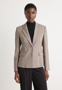 Beige přizpůsobený blazer s jemnou texturou, zapínání na jeden zlatý knoflík, klopové límce a přední kapsy. Nositý přes černý rolák.
