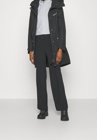 Giacca impermeabile nera con cappuccio sopra un pile grigio chiaro con cerniera, abbinata a pantaloni neri a vita alta e scarpe da outdoor scure.