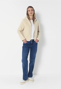 Giacca in pile beige con cappuccio, design con zip frontale, abbinata a jeans blu a gamba dritta e sneakers chiare. Look semplice e casual.