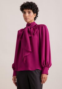 Blusa fucsia con un grande fiocco al collo, spalle plissettate e maniche voluminosi. Tessuto morbido e fluido; texture liscia.