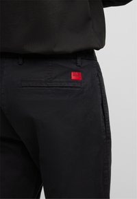 Sorte bukser med en rød logo-patch. Glat stof, lige snit, med en baglomme og diskrete sømdetaljer.