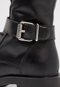 Bottine en cuir noir avec boucle en métal argenté et détail de bride sur le dessus, finition lisse et texturée.
