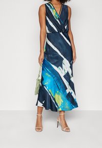 Volná, bezrukávová maxi šaty v námořnické modři s výraznými bílými a zelenými štětcovými vzory. Hladká tkanina s staženým pasem, doplněné o páskové podpatky.