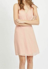 Žena s vlnitými vlasy má na sobě bezramínkovou světle růžovou šaty ke kolenům a zkřížené ruce před bílým pozadím.