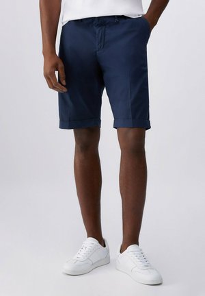 Uomo che indossa pantaloncini blu navy con orlo risvoltato e sneakers bianche, in piedi con una mano in tasca contro uno sfondo uniforme.