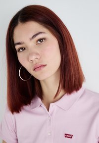 Camisa polo de color rosa claro con cuello, que presenta un pequeño logotipo rojo de Levi's. Cabello lacio de color castaño rojizo hasta los hombros; grandes pendientes de aro plateados.