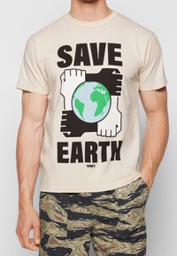 Beige tričko s nápisem "ZACHRAŇTE ZEMI", čtyři propletené ruce v černé a bílé kolem ilustrace zelené a modré Země.
