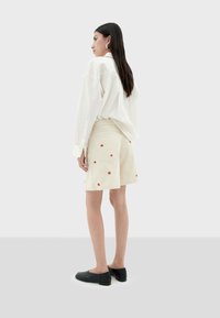Witte oversized blouse met lange mouwen, gecombineerd met een beige rok met rode bloemenborduursels, en zwarte platte schoenen.