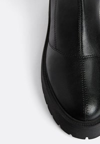 Černá kožená kotníková bota s zaoblenou špičkou, hladkým povrchem a silnou gumovou podrážkou. Zesílené švy podél svršku.